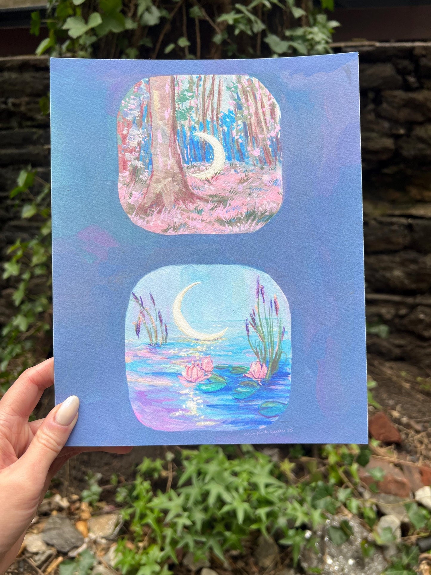 May include: A watercolor painting on blue paper featuring two square vignettes. The top vignette depicts a forest scene with a crescent moon. The bottom vignette shows a moonlit pond with water lilies. The painting is held by a person.